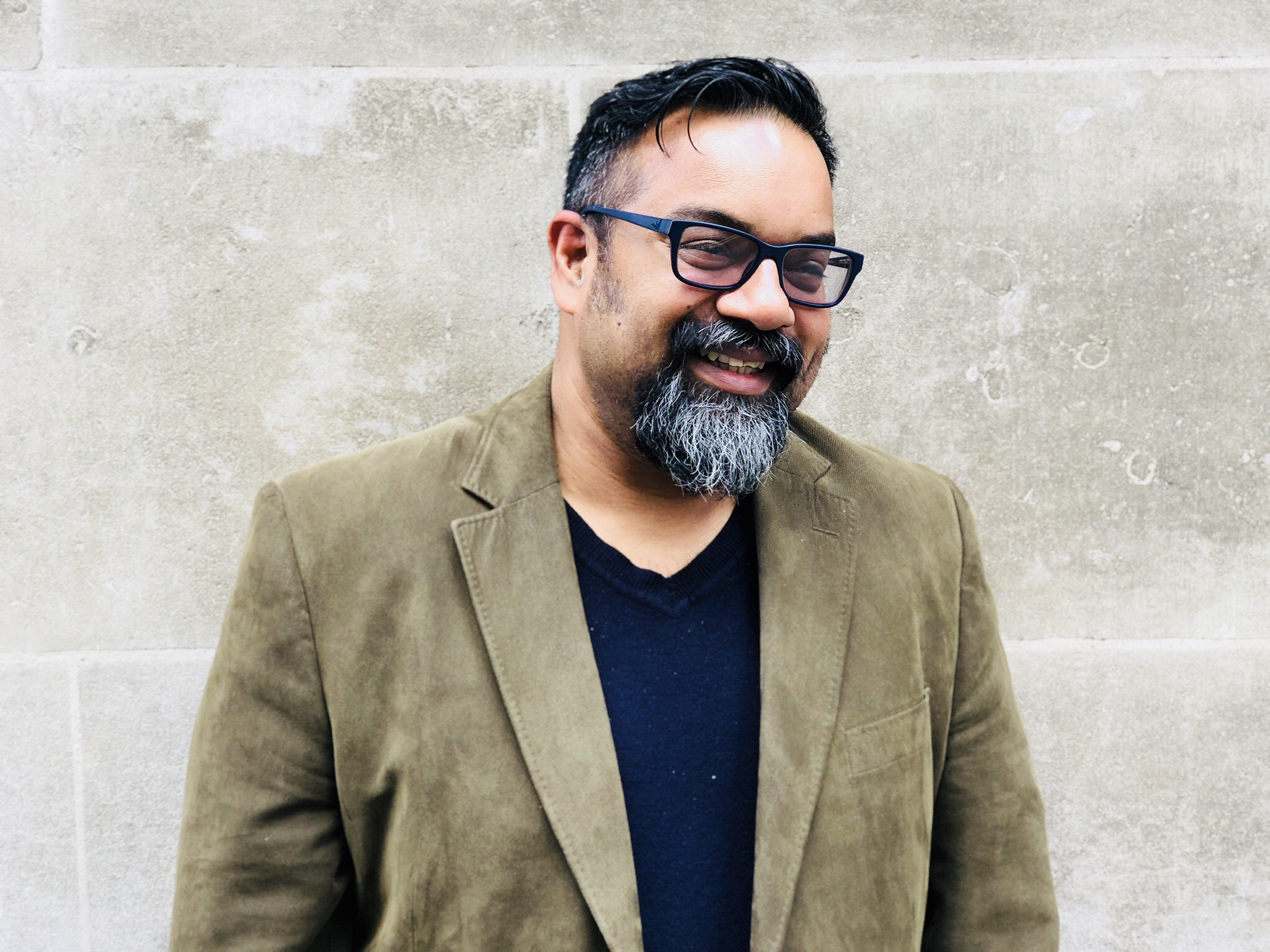 Atif Choudhury laughing, wearing an olive blazer, against a concrete wall